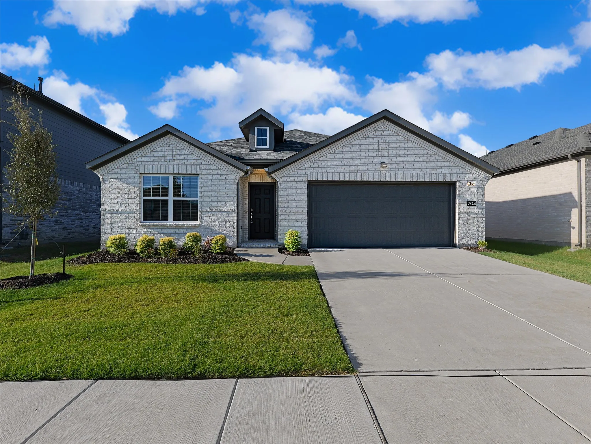 Charming brick residence featuring a modern black two-car garage, arched roofline with decorative dormers, manicured front lawn with established landscaping, concrete driveway, and welcoming entry with dark wood-finish door and transom window