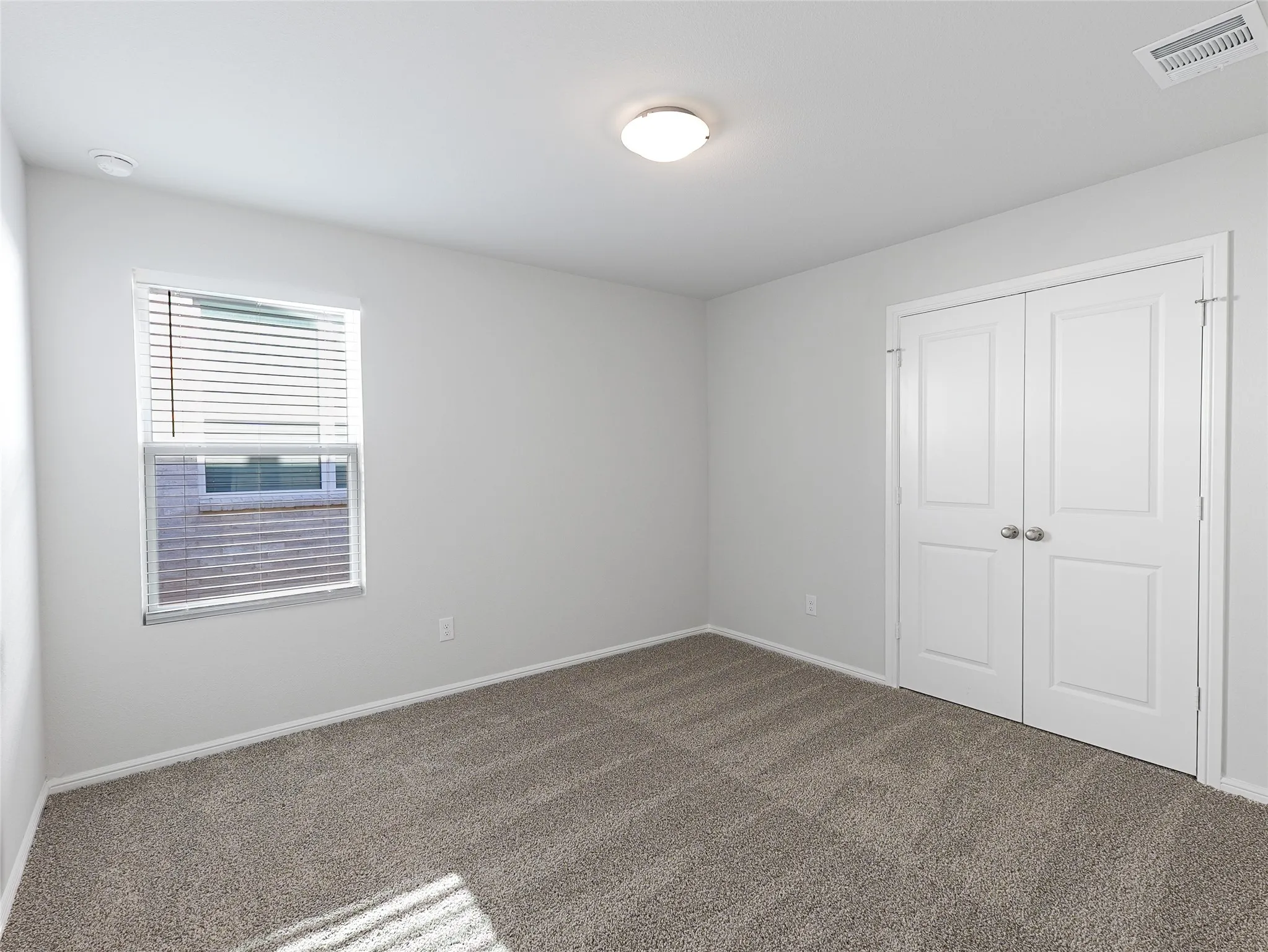 Bright secondary bedroom showcasing neutral wall finishes, carpeted flooring, ample natural light from a multi-pane window with horizontal blinds, a recessed ceiling light fixture, and a spacious double-door closet with white six-panel doors