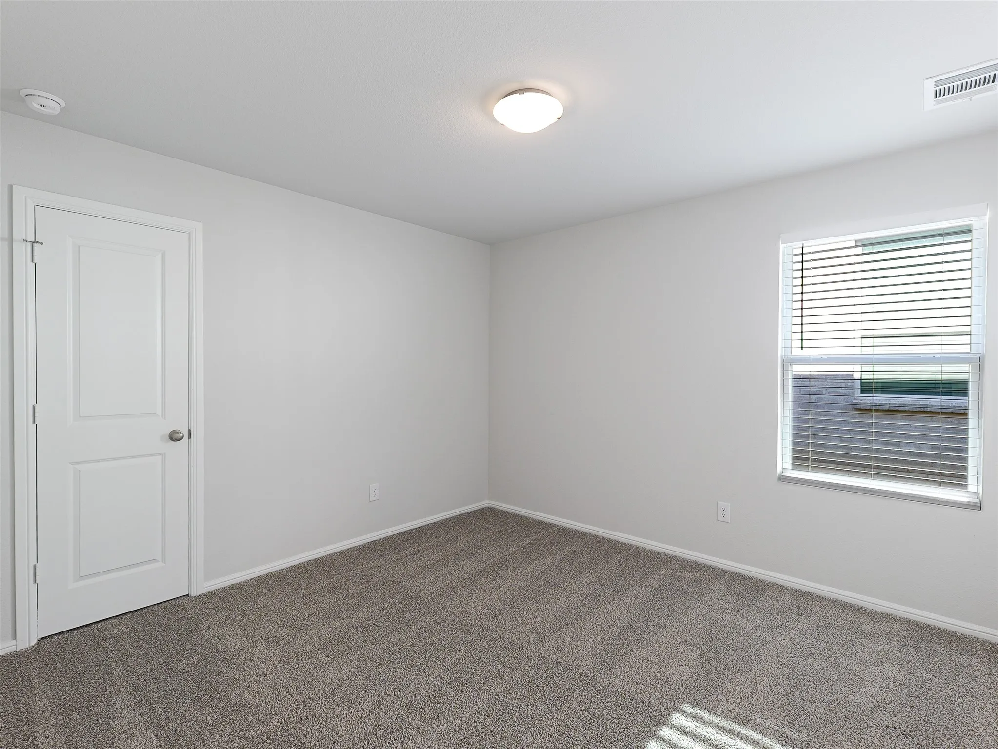 Bright secondary bedroom showcasing neutral wall finishes, plush carpeting, a single window with horizontal blinds providing natural light, and a white six-panel entry door