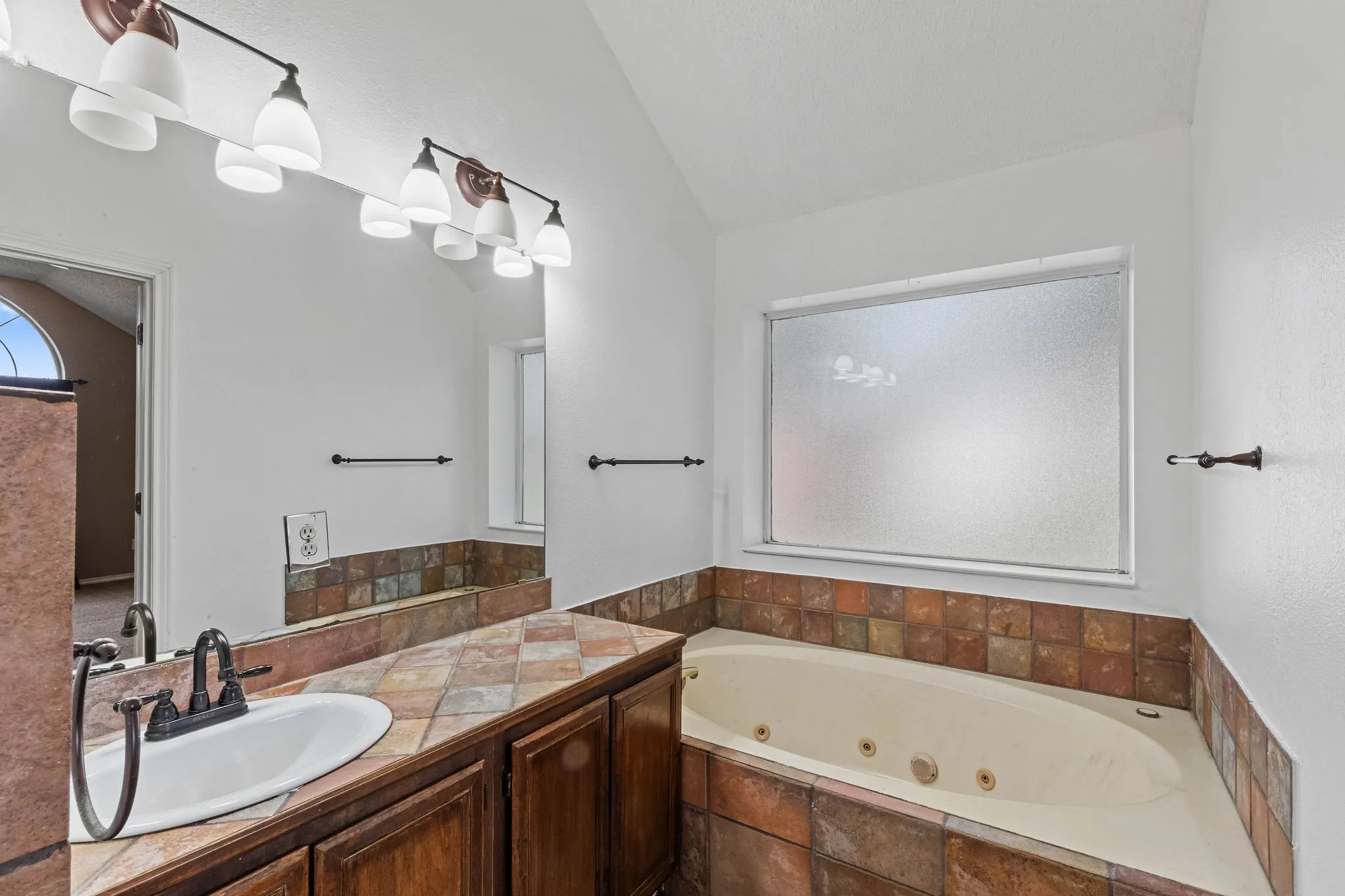Corner soaking tub with tiled surround, wood-finish vanity with an integrated sink, and overhead vanity lighting
