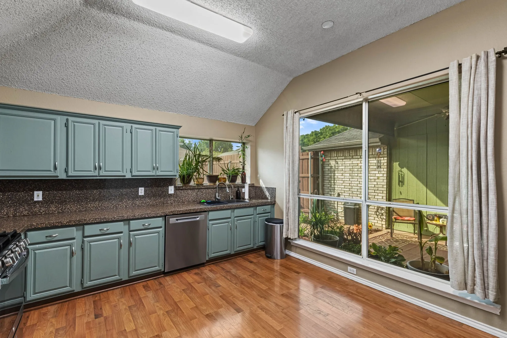 Vaulted ceiling, wood-finish flooring, full-height window overlooking an outdoor patio, teal cabinetry, and a built-in stainless steel dishwasher