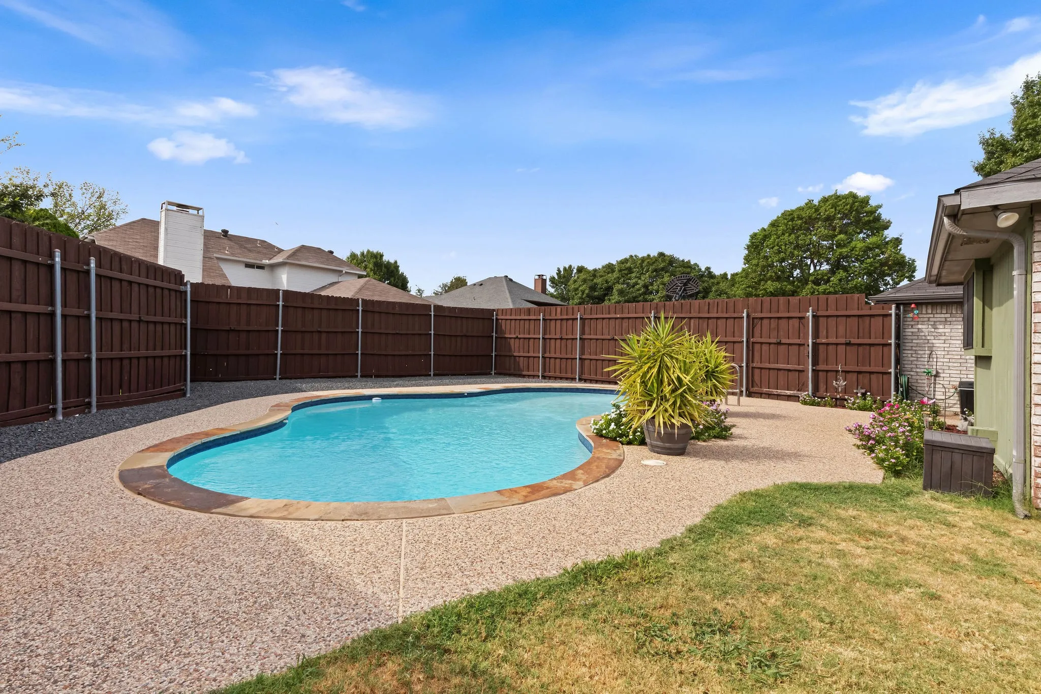 Curved swimming pool with stone coping, surrounded by an exposed aggregate patio, and a privacy fence