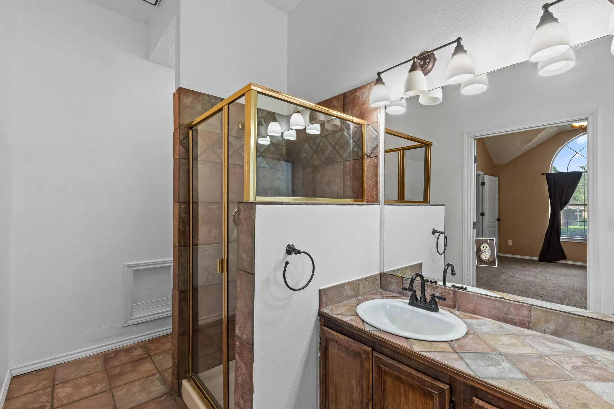 Enclosed glass shower stall with gold-tone framing, a vanity with a drop-in sink, a tile countertop, and a multi-bulb vanity light fixture