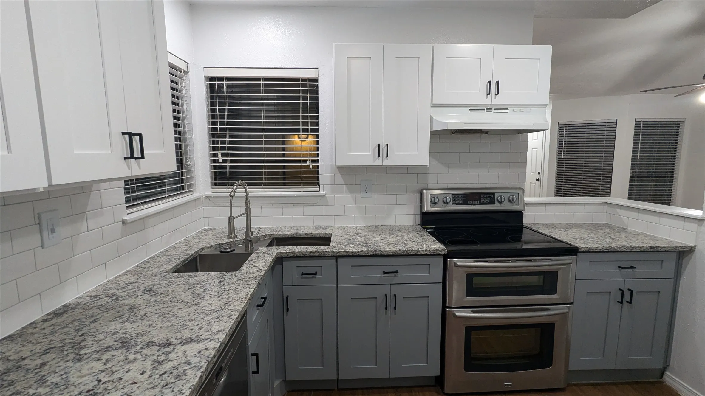 Kitchen featuring light-toned stone countertops, white subway tile backsplash, stainless steel sink with gooseneck faucet, two-tone cabinetry with black hardware, and a stainless steel double oven range