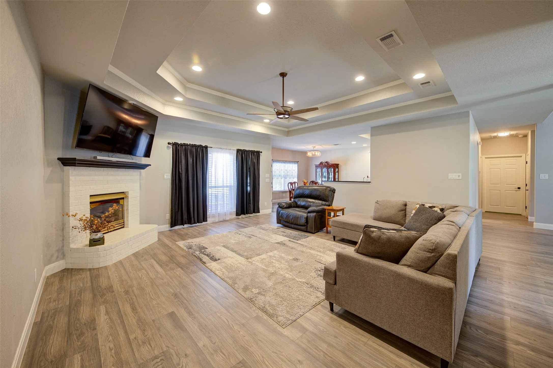 Wood-finish flooring throughout, tray ceiling with recessed lighting, ceiling fan, and a fireplace with a dark wood mantel