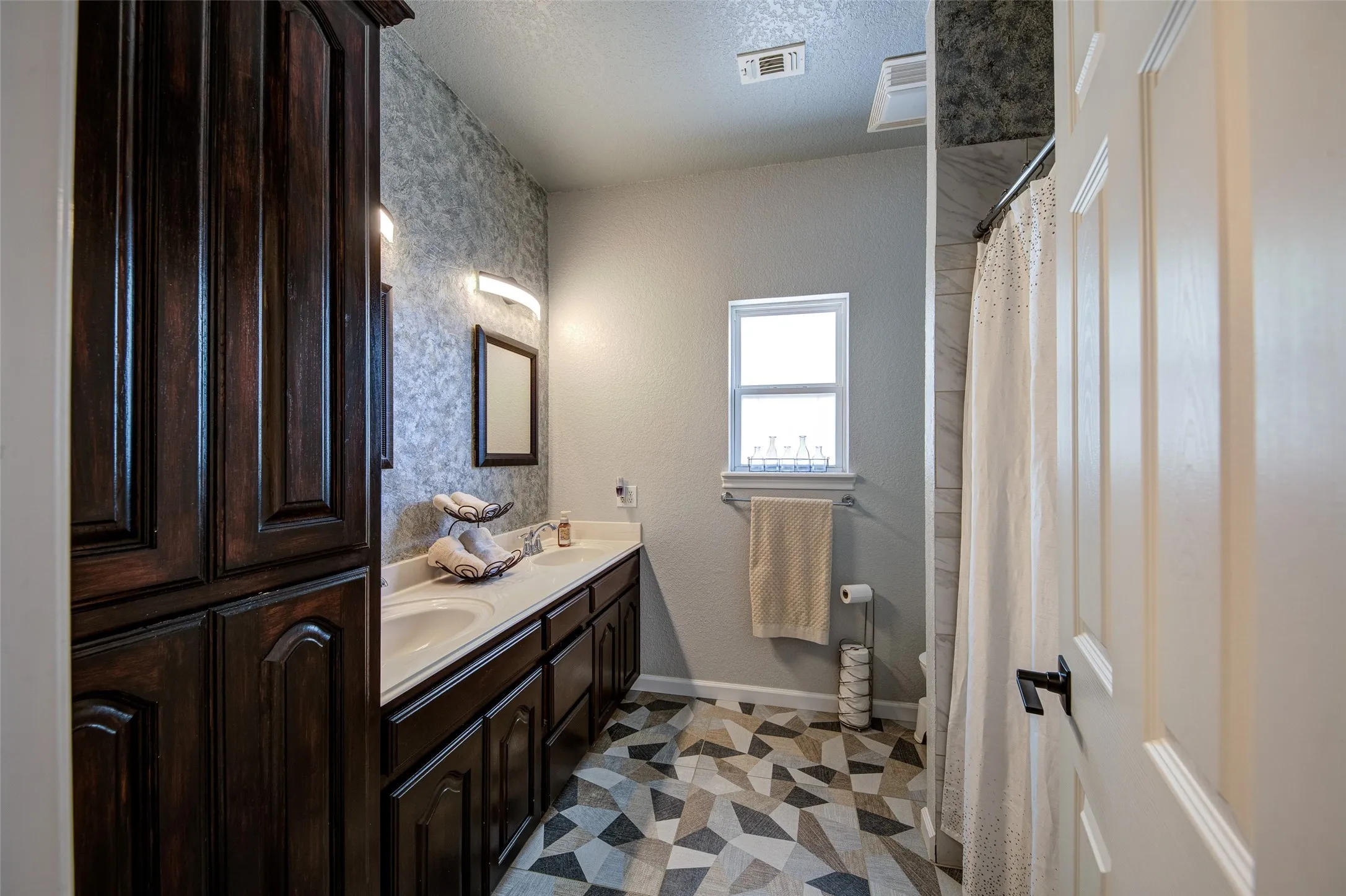 Dual vanity with undermount sinks, extended countertop, built-in storage cabinetry, geometric pattern flooring, and window for natural light