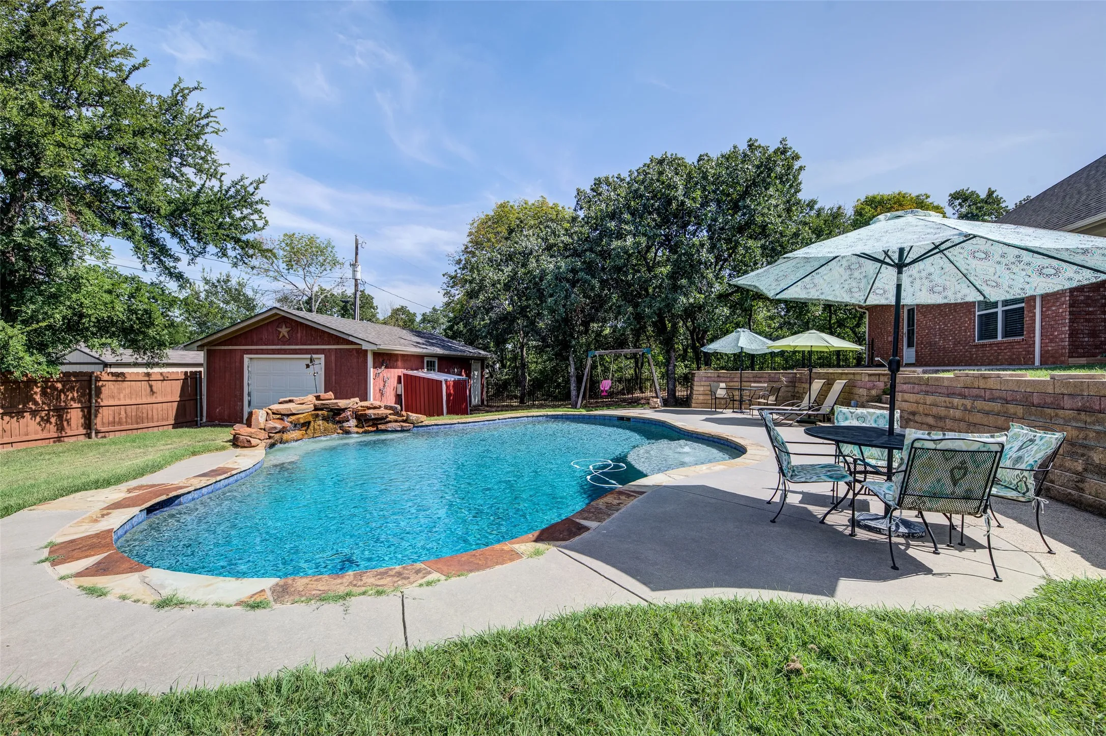 Resort-style backyard oasis featuring a custom-shaped saltwater pool with sun shelf, expansive concrete decking, mature landscaping with established trees, and adjacent brick patio with pergola shade structure