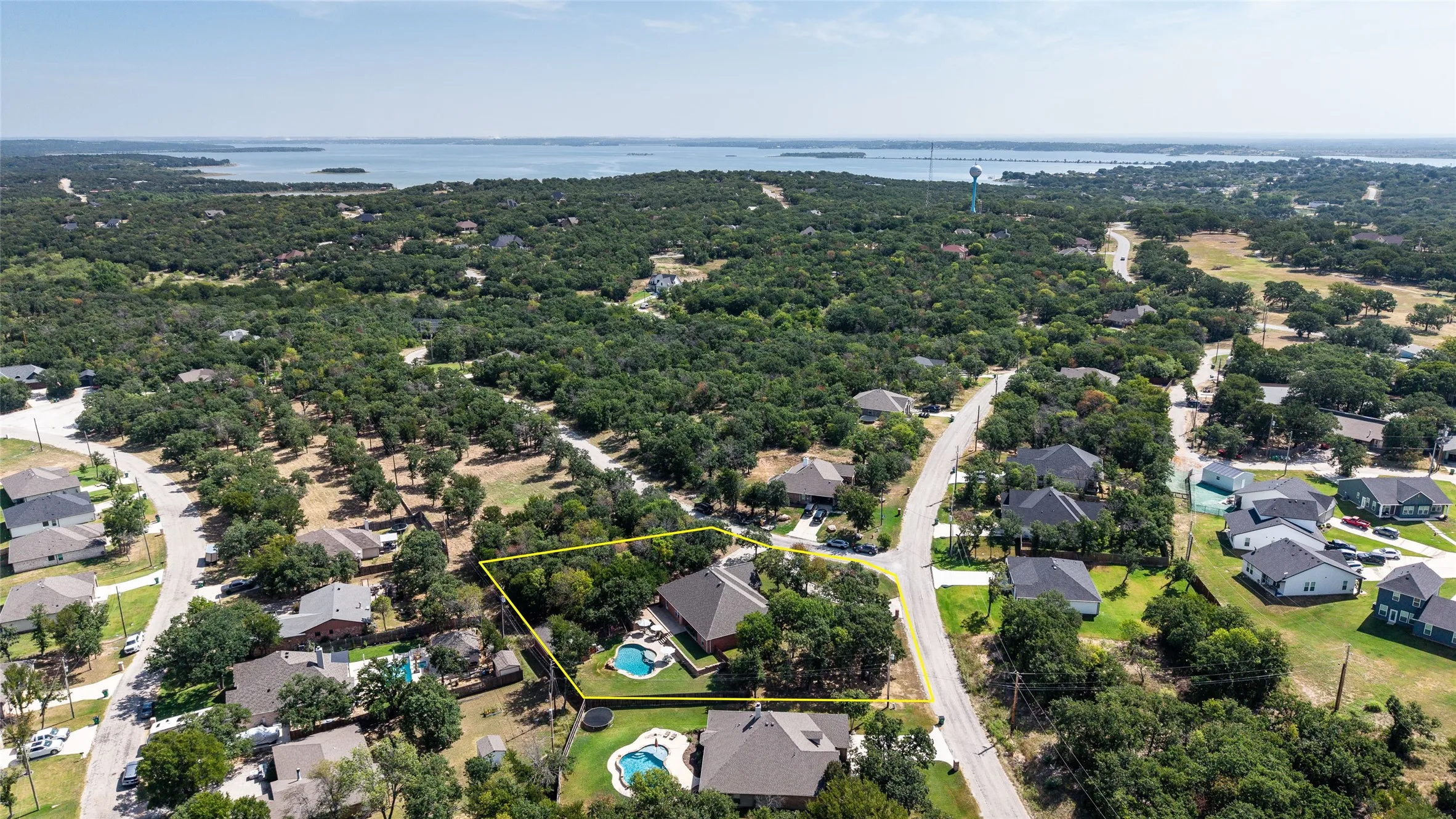 Aerial perspective showcasing a residential property with an in-ground swimming pool, surrounded by mature trees, and a distant body of water