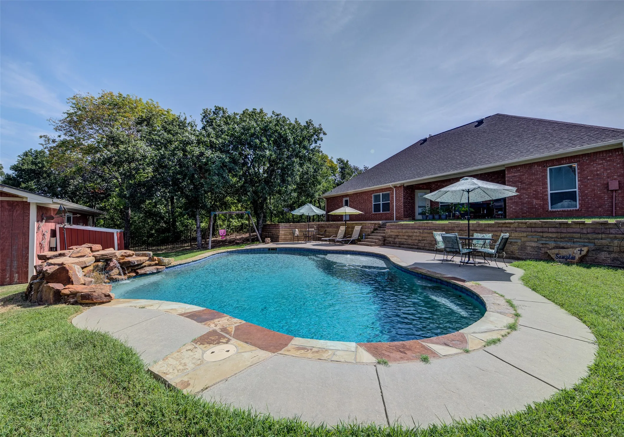 Curved swimming pool with an integrated rock waterfall feature, concrete and flagstone surround, and an elevated patio area