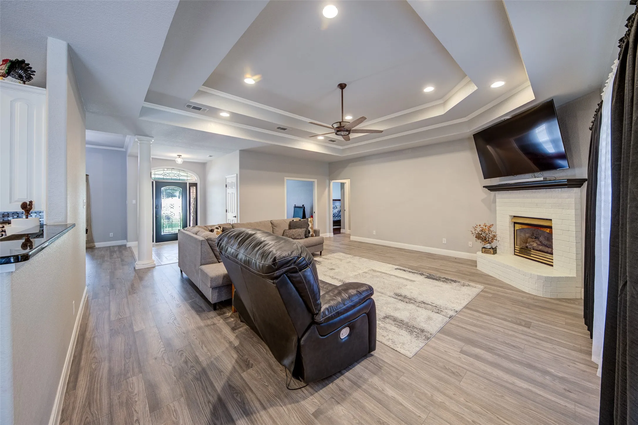 Spacious living area featuring wood-finish flooring, a tray ceiling with recessed lighting, a ceiling fan, and a fireplace with a white brick surround