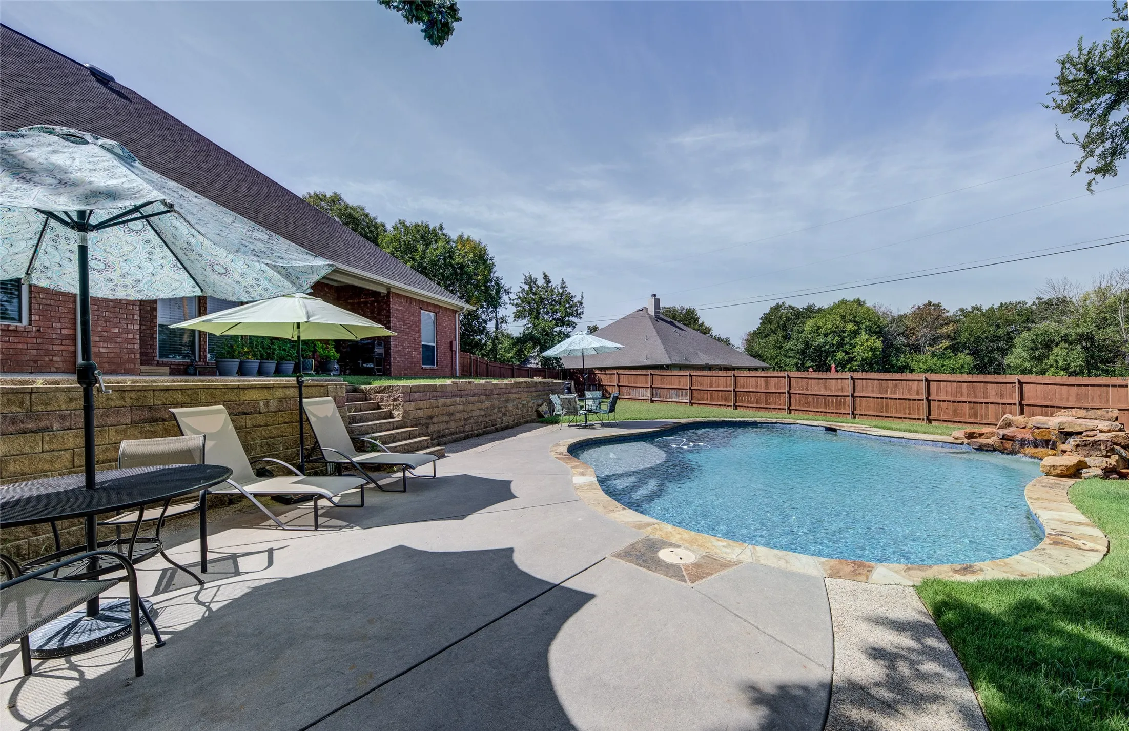 Inviting backyard oasis featuring a freeform swimming pool with rock waterfall, extensive concrete patio, tiered retaining wall with steps, and a privacy fence