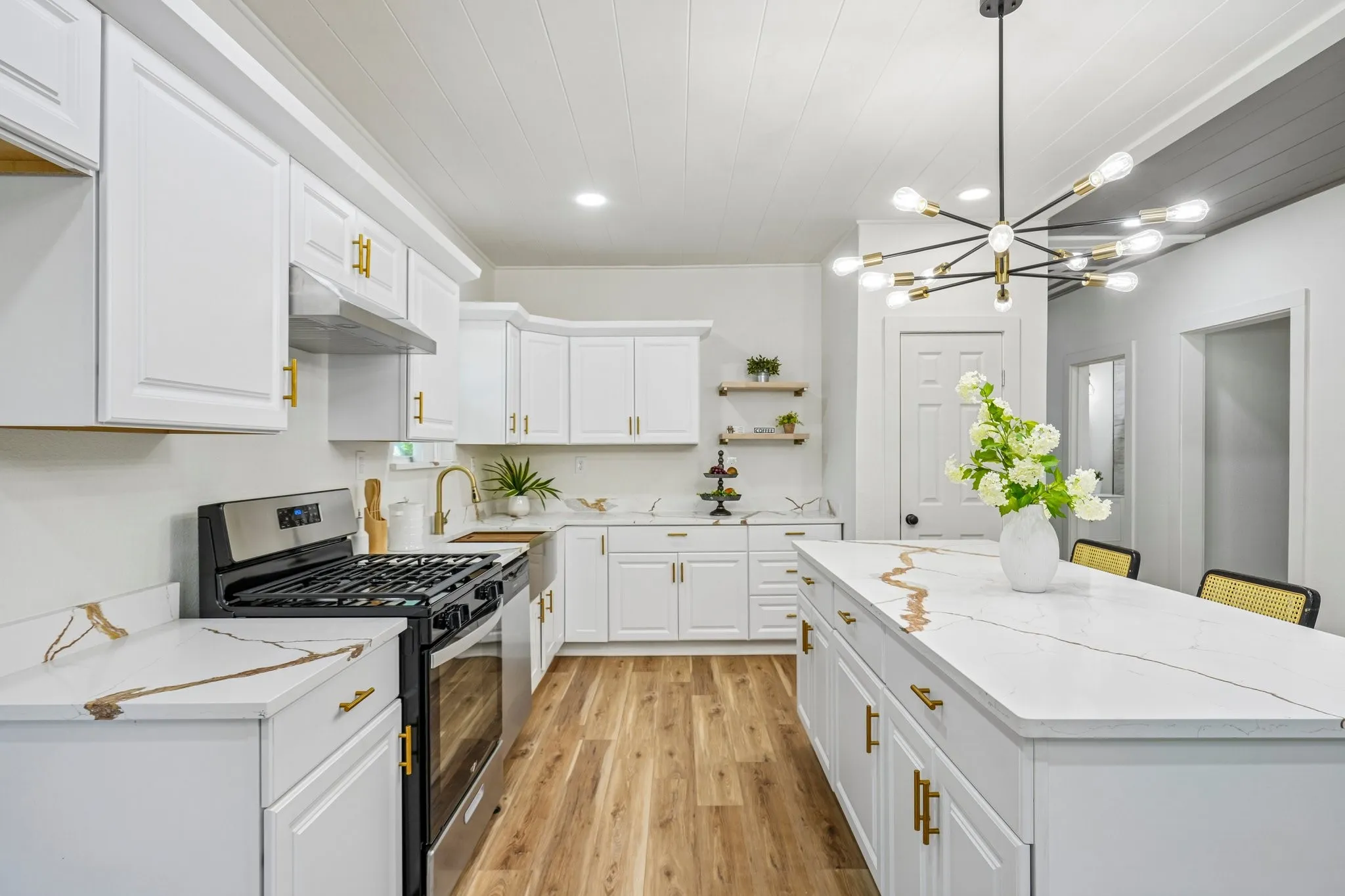 White shaker cabinetry with gold hardware, stainless steel range, white countertops with gold veining, wood-finish flooring, and a modern sputnik chandelier