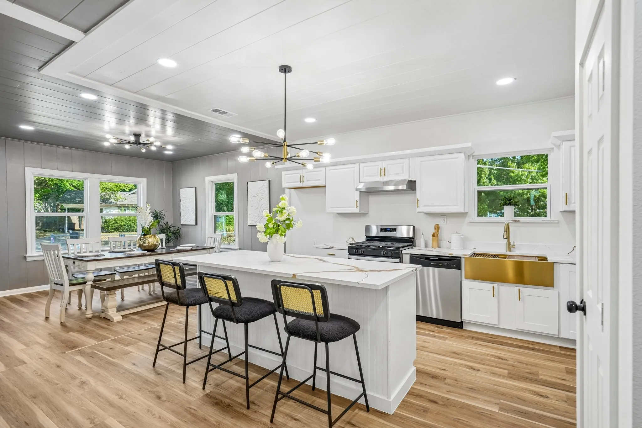 Open-concept kitchen and dining area featuring wood-finish flooring, white cabinetry, a gold-tone apron sink, stainless steel appliances, and a central island with a light-toned countertop