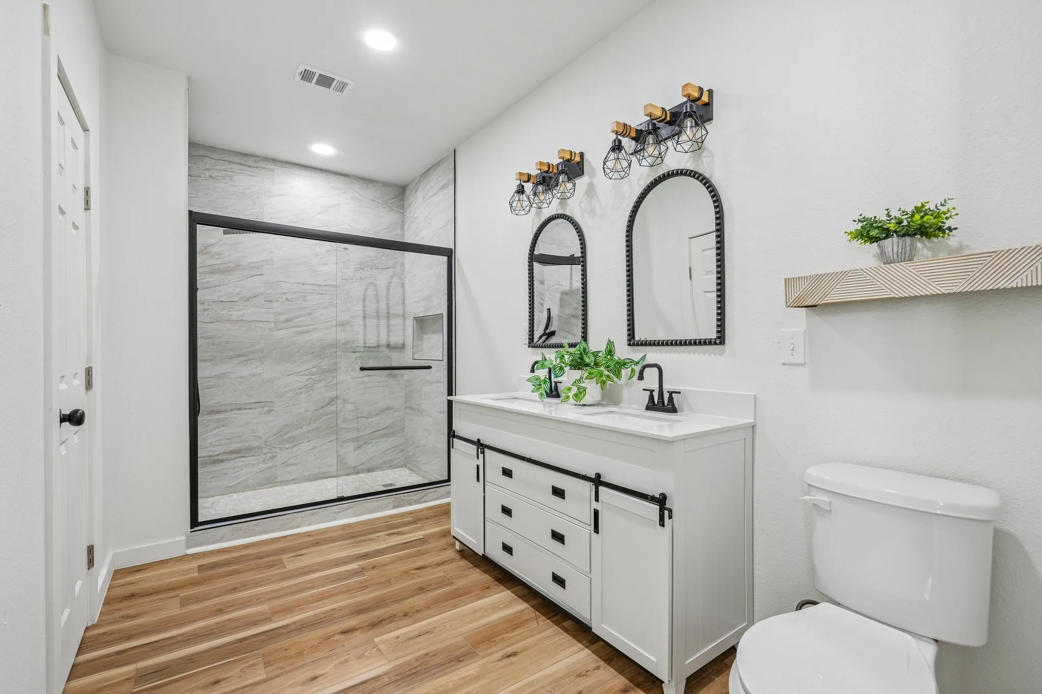 Dual vanity with undermount sinks, wood-finish flooring, tiled shower with glass enclosure, white cabinetry with black hardware, and overhead recessed lighting