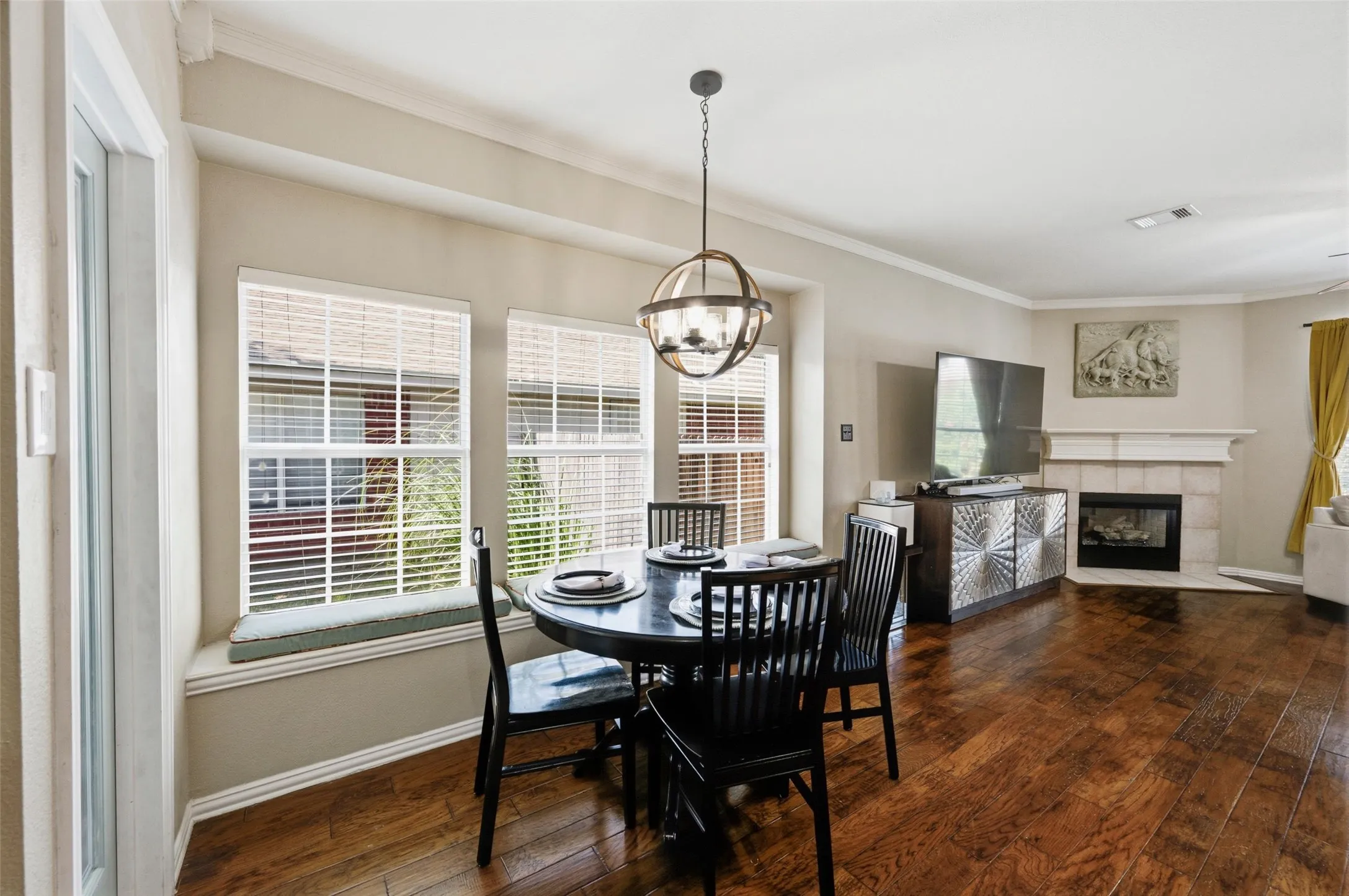 Hardwood-finish flooring throughout, three windows with blinds, built-in window seating, fireplace with tile surround, and crown molding