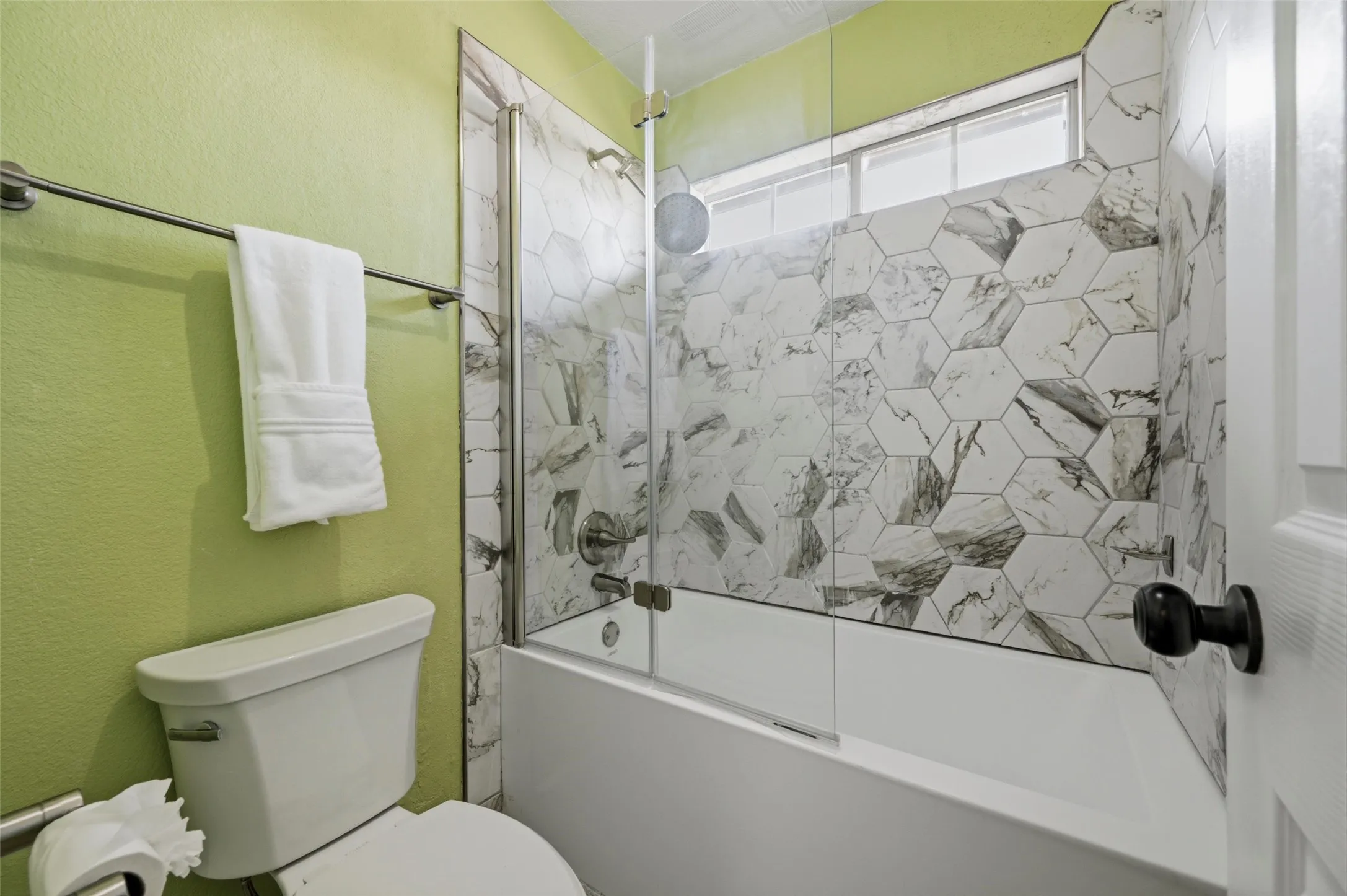 Bathtub with a glass shower enclosure, white hexagonal wall tile, and a privacy window