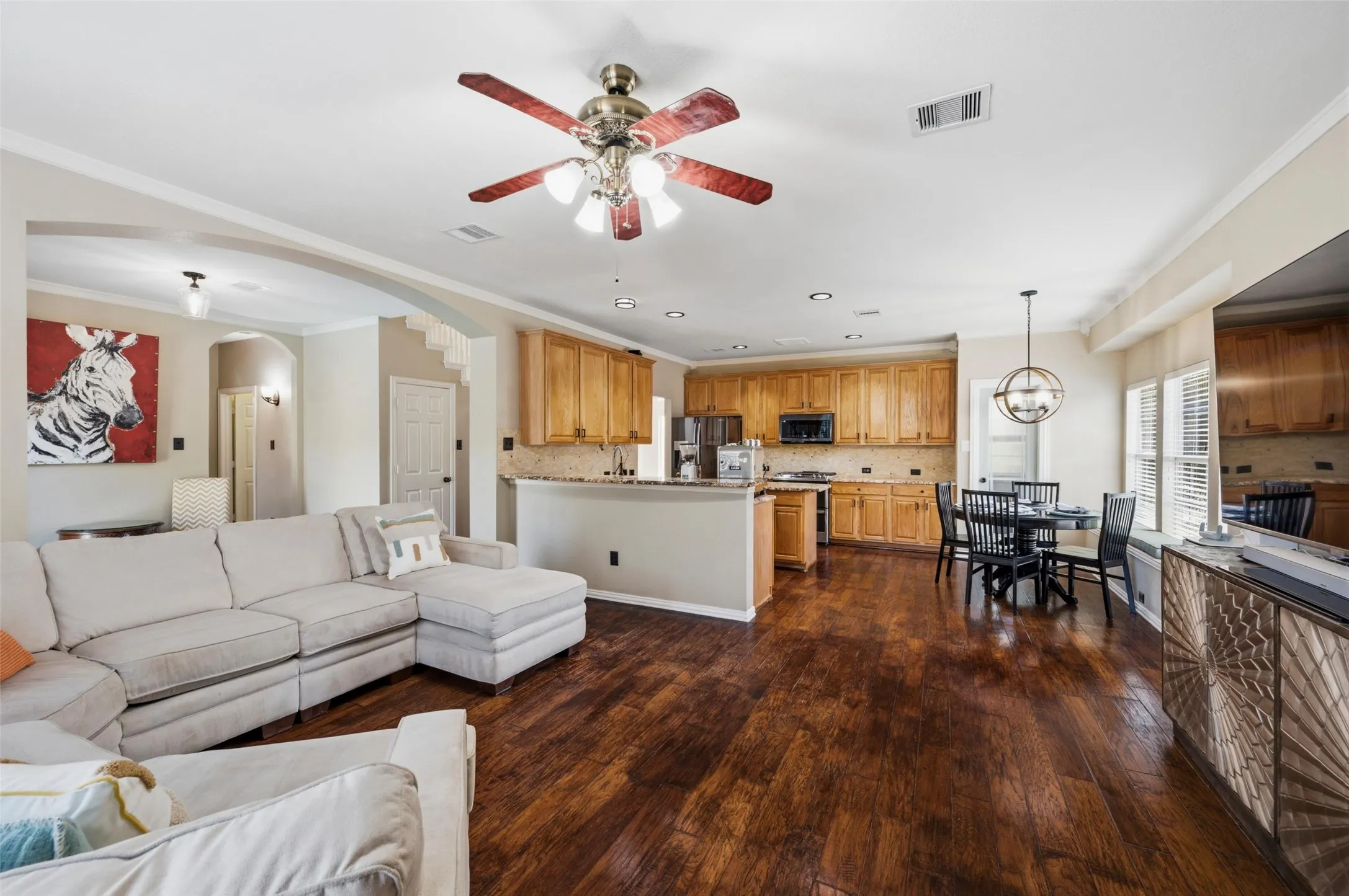 Open-concept living space featuring wood-finish flooring, a kitchen with wood cabinetry and a tile backsplash, and a dining area with a circular pendant light fixture