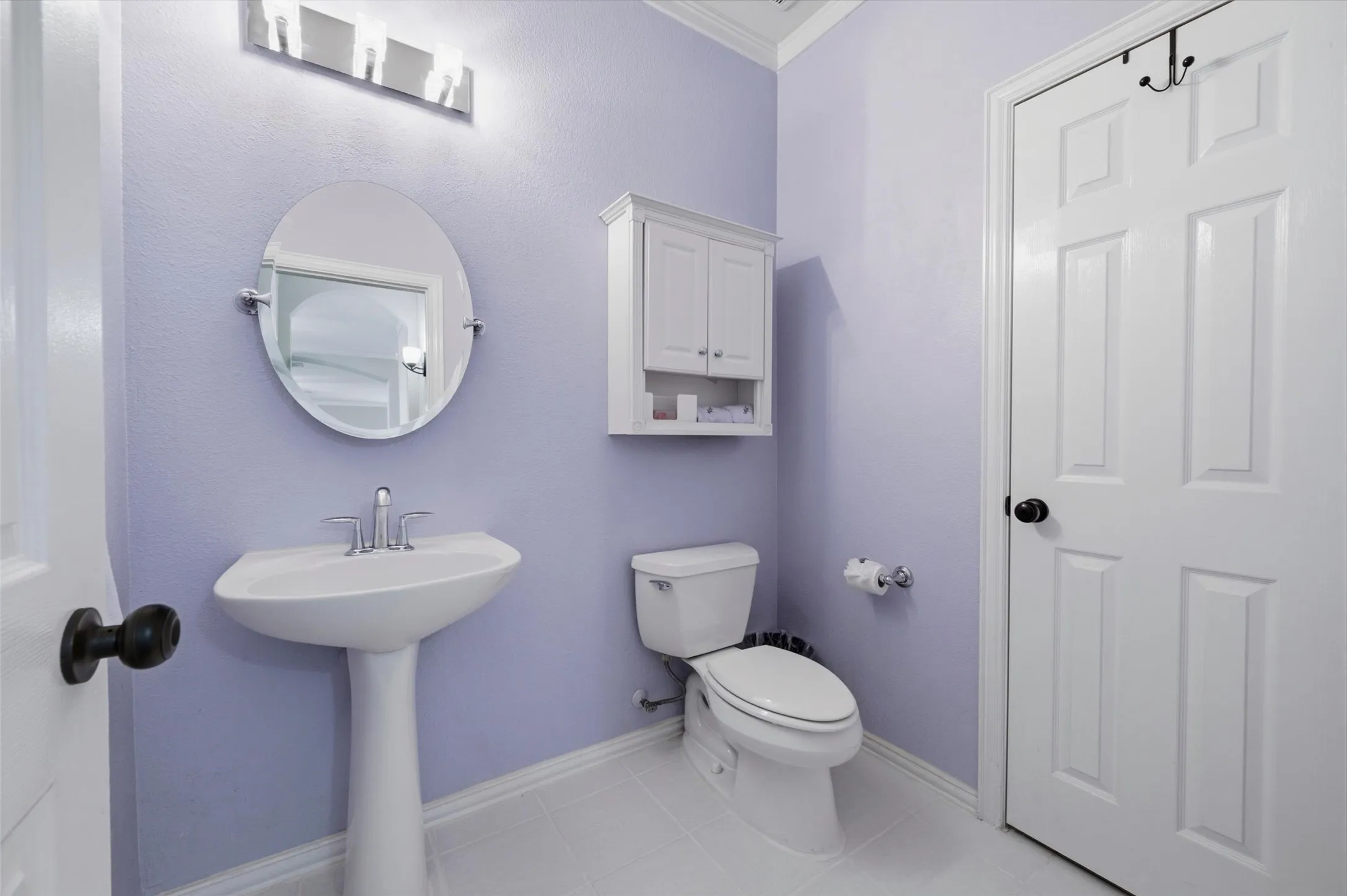 Pedestal sink with chrome faucet, wall-mounted mirror, overhead vanity lighting, white wall cabinet, and white paneled door