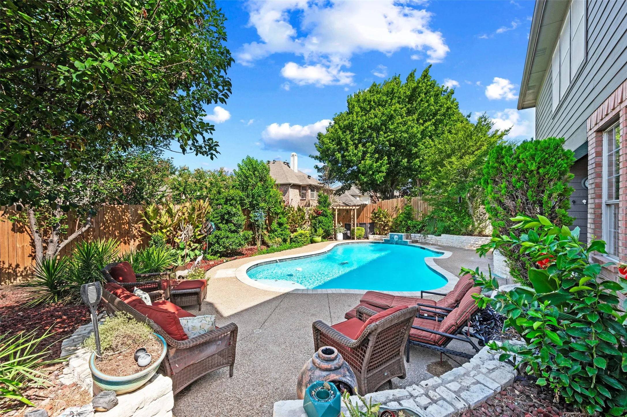 Curved swimming pool with an exposed aggregate deck, surrounded by mature landscaping, a wooden privacy fence, and an exterior brick and siding facade