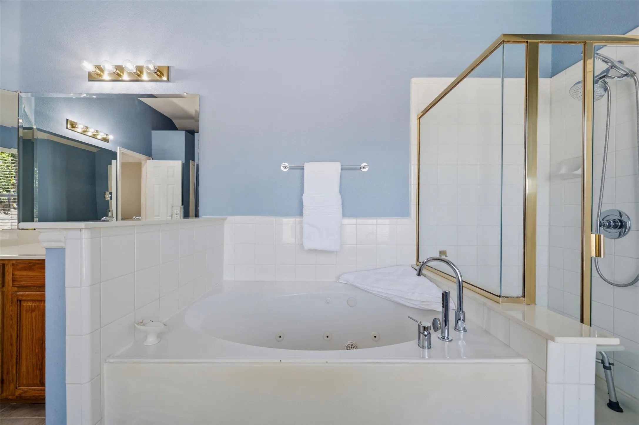 Built-in jetted tub, tiled surround, glass-enclosed shower with gold-tone framing, vanity with wood-finish cabinetry, and mirrored wall with overhead vanity lighting