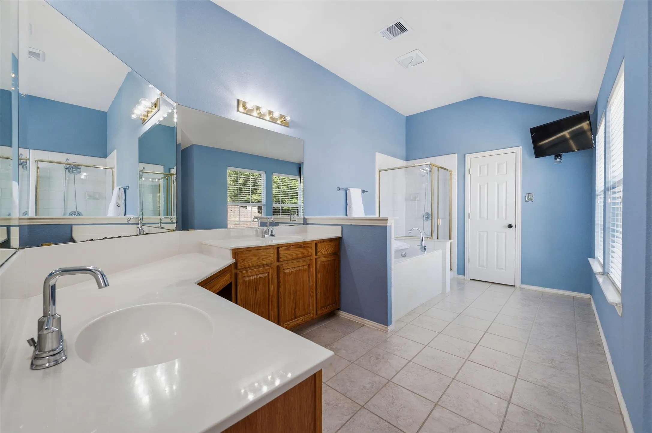 Vaulted ceiling, dual vanity with wood cabinetry and light-toned countertops, tiled flooring, glass-enclosed shower, and built-in tub