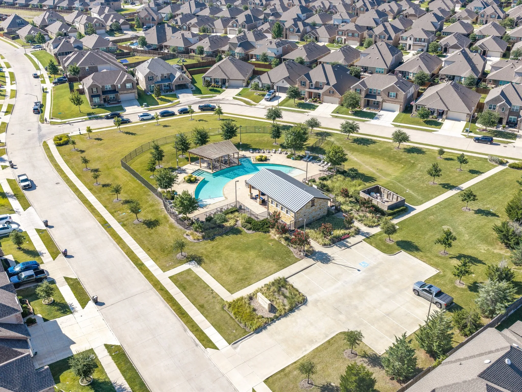 Community pool with surrounding lounge areas, stone-clad building with metal roof, open-air pavilion, and adjacent green space with walking paths