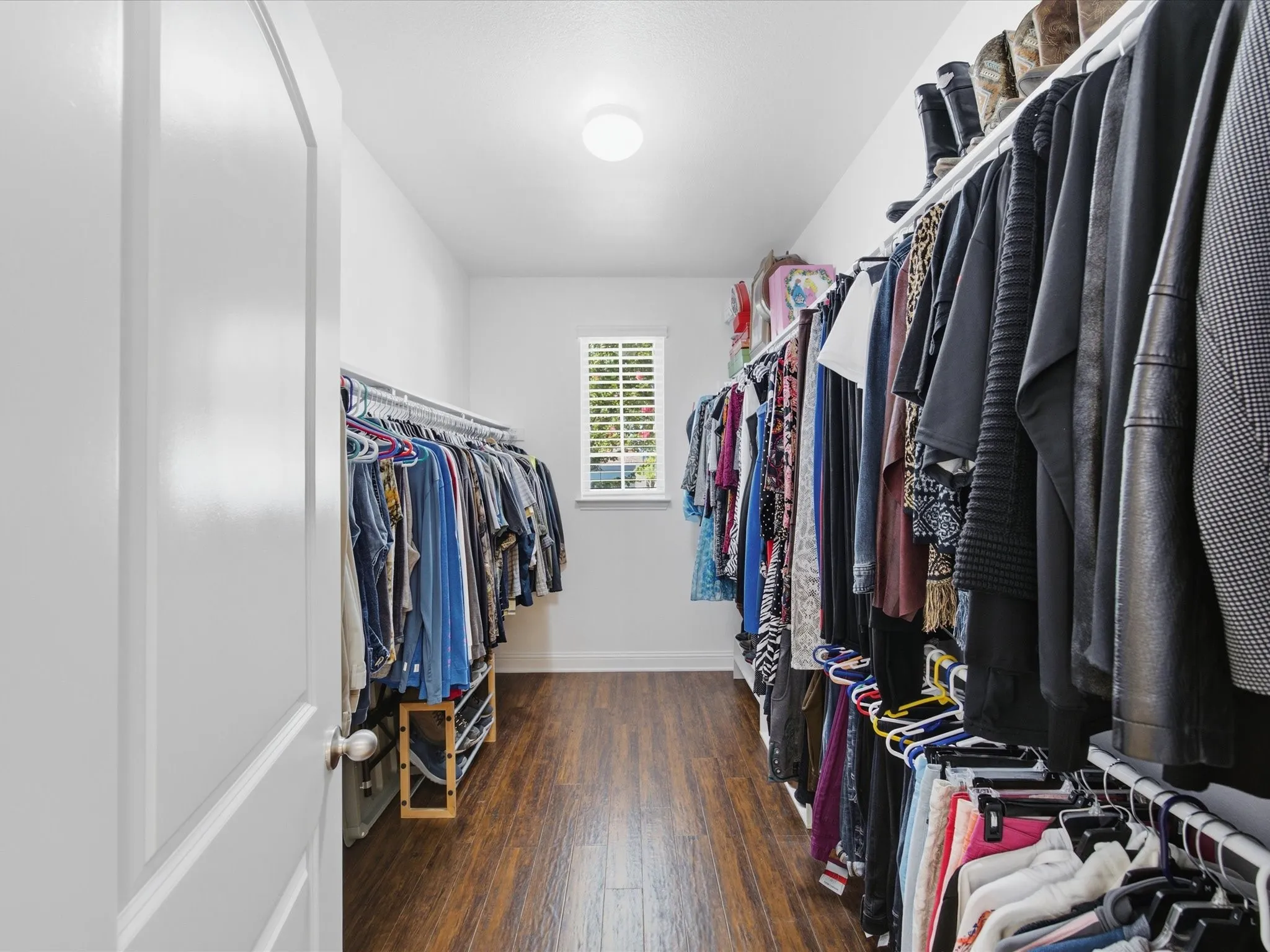 Walk-in closet featuring wood-finish flooring, a window with horizontal blinds, and multiple clothing racks