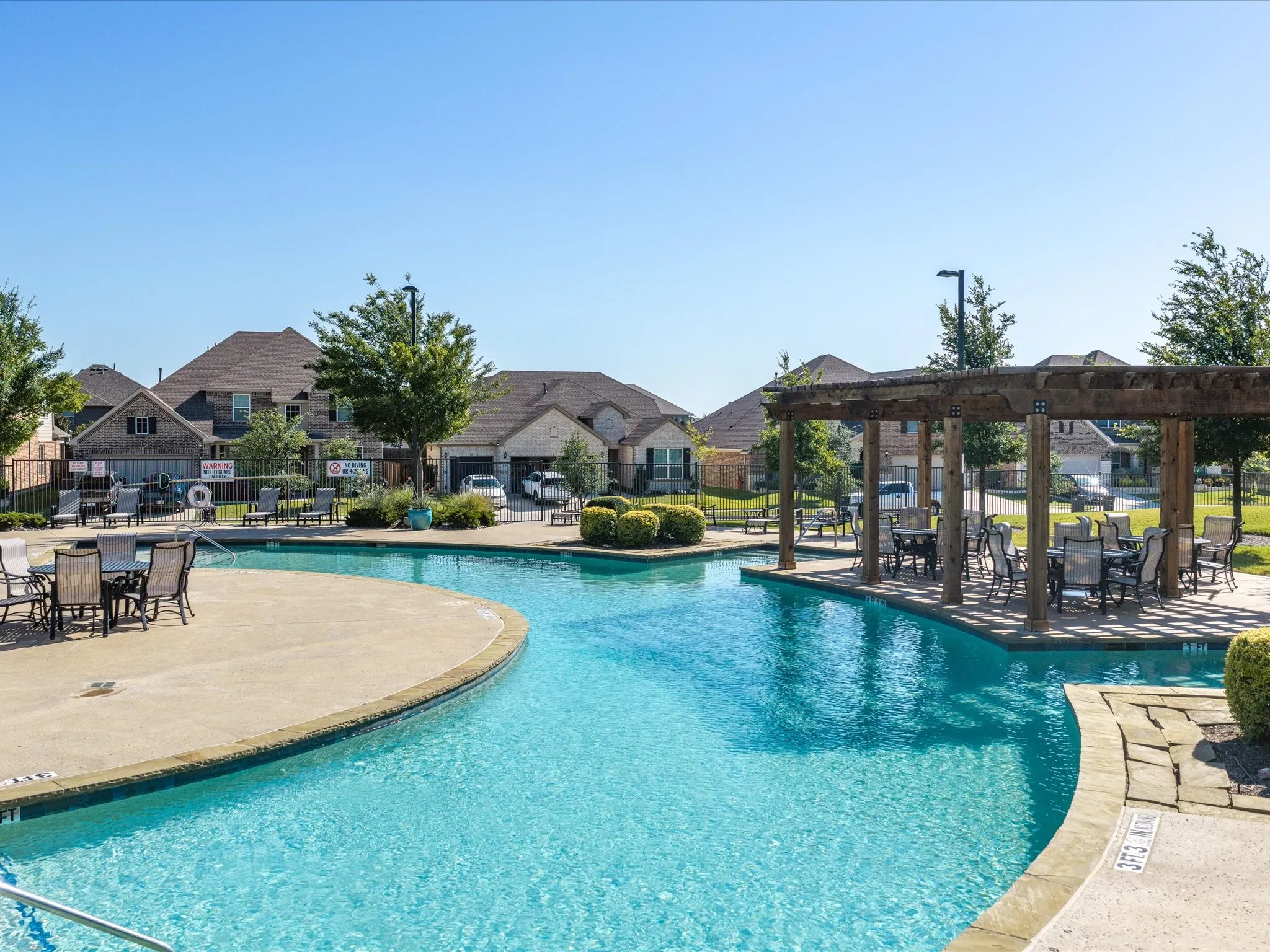 Curved-edge swimming pool with a surrounding concrete deck, adjacent to a wooden pergola with seating, and landscaped green areas