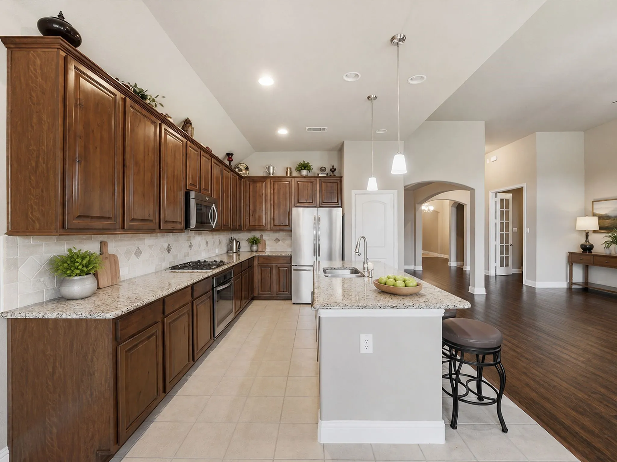Spacious kitchen featuring extensive wood cabinetry, light-toned granite countertops, stainless steel appliances, and a central island with pendant lighting