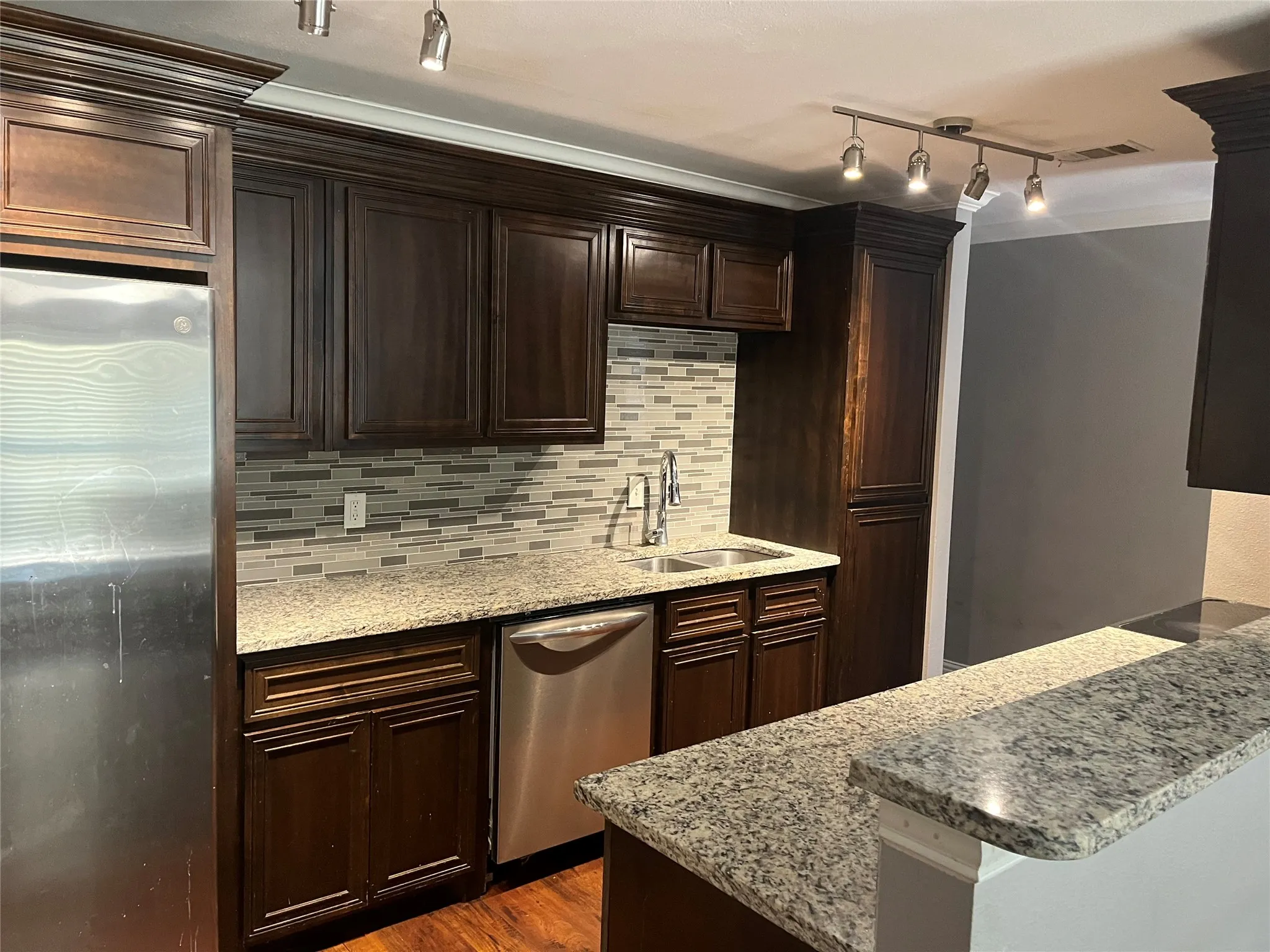 Dark wood-finish cabinetry with crown molding, stainless steel appliances, granite countertops, mosaic tile backsplash, and wood-finish flooring