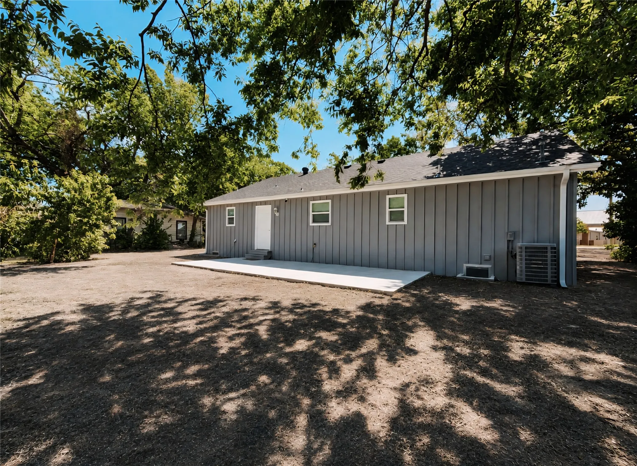Spacious rear yard with a newly poured concrete patio, gray board and batten siding, white exterior door, and white frame windows