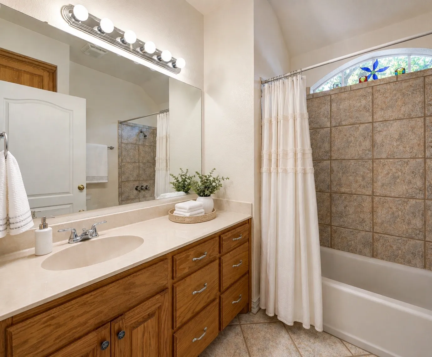 Vaulted ceiling bathroom featuring a single vanity with wood-finish cabinetry, a large mirror, a bath-shower combination with tile surround, and an arched window