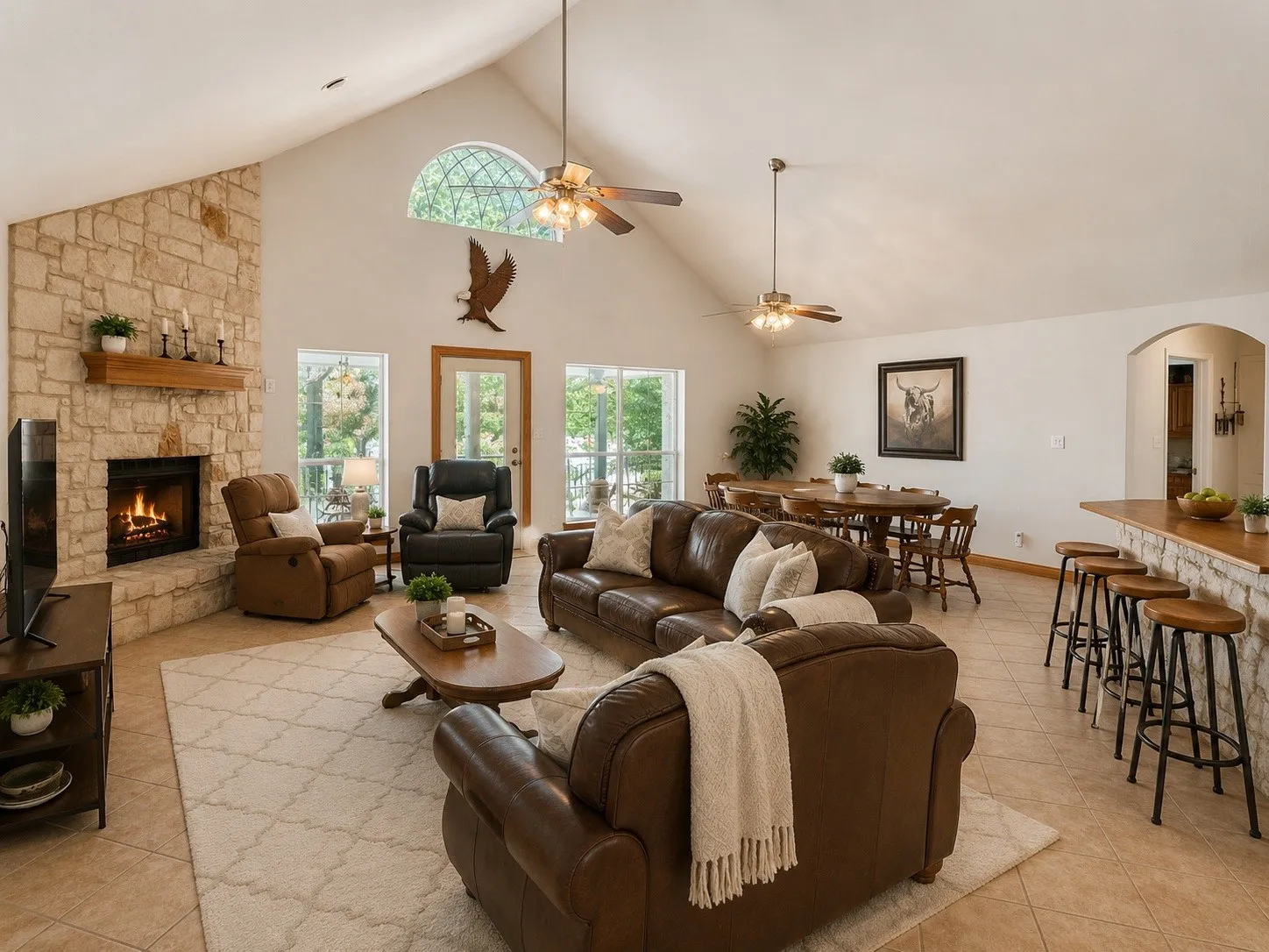 Vaulted ceiling, stone fireplace with wood mantel, arched window, ceiling fans, and tiled flooring