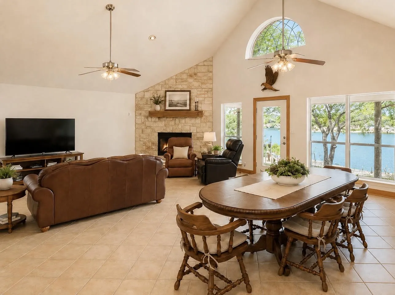 Vaulted ceiling with dual ceiling fans, stone-clad fireplace with wood mantle, tile flooring, and large windows with water scenery