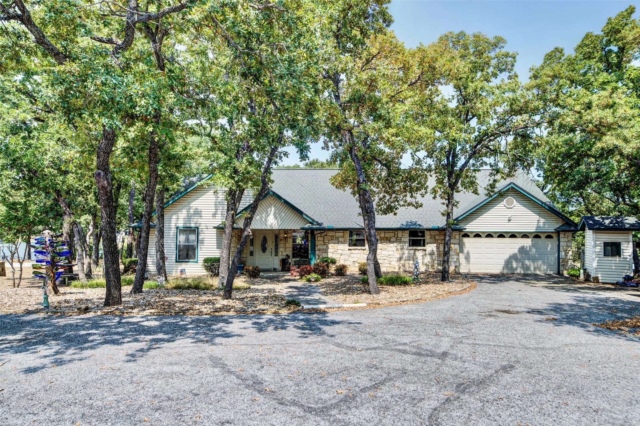 Stone and siding exterior with a multi-car garage, mature trees, and a gravel driveway