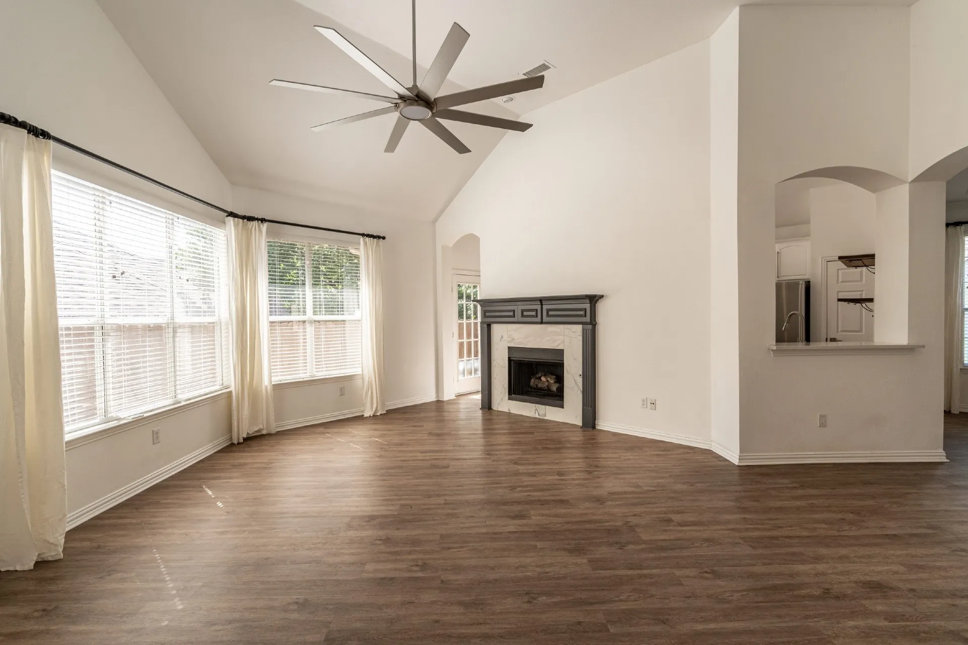 Vaulted ceiling with a large ceiling fan, wood-finish flooring, multiple windows, a fireplace with a mantel, and an arched pass-through window