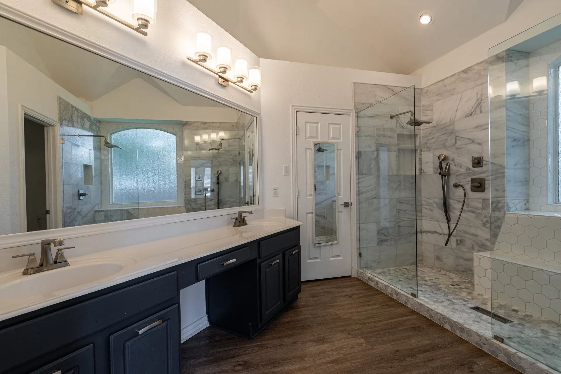 Dual vanity with white countertops, expansive mirror with vanity lighting, glass-enclosed shower with tiled walls and a built-in bench, wood-finish flooring