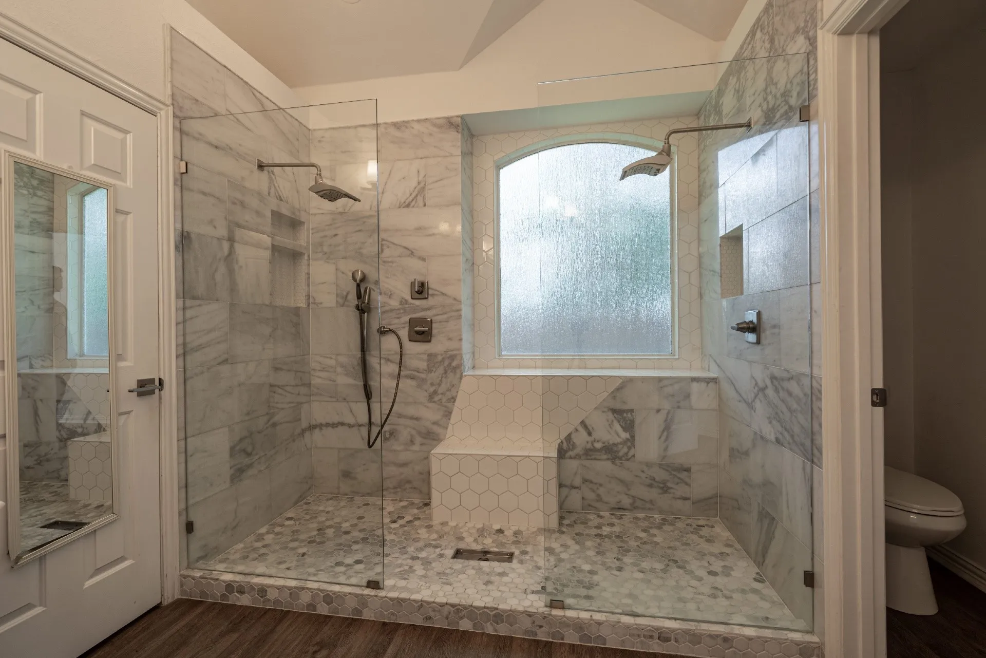 Glass-enclosed shower with a vaulted ceiling, dual shower heads, a built-in bench, and an arched privacy window