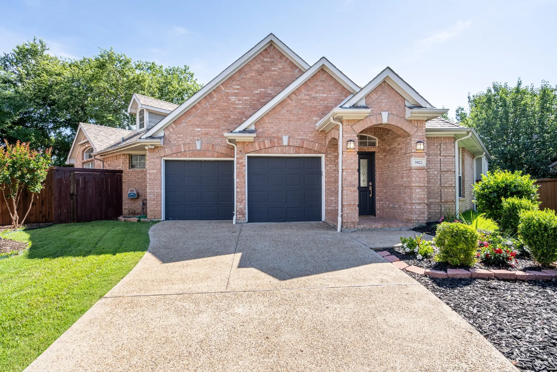 Brick exterior with a gabled roofline, two-car garage, arched entry, and mature landscaping