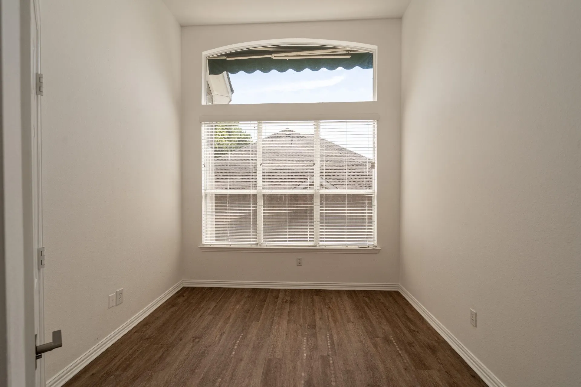 Arched transom window with a green awning and rectangular window featuring blinds, wood-finish flooring, and white walls