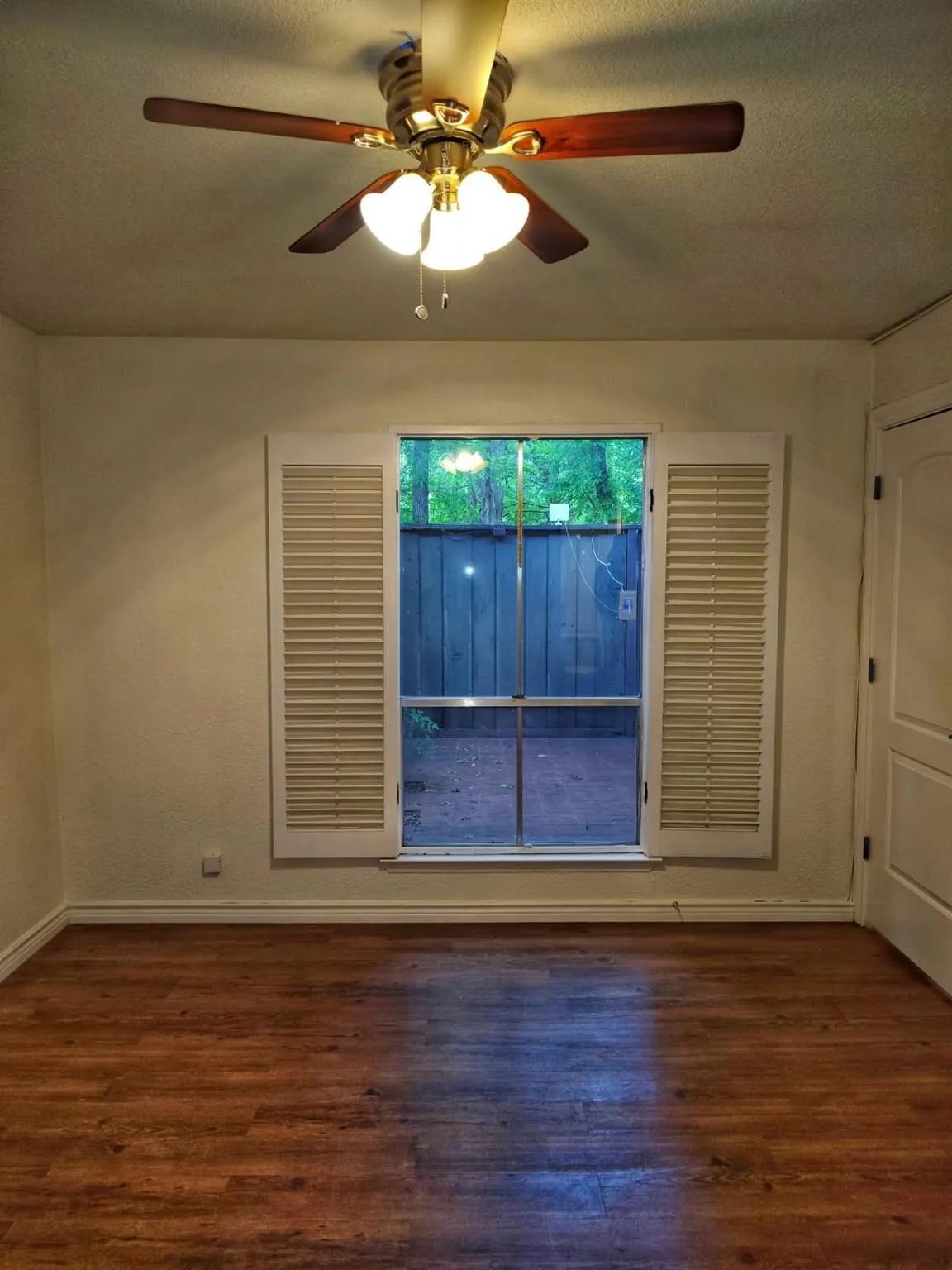 Wood-finish flooring, ceiling fan with integrated lighting, window with louvered shutters, and a paneled interior door