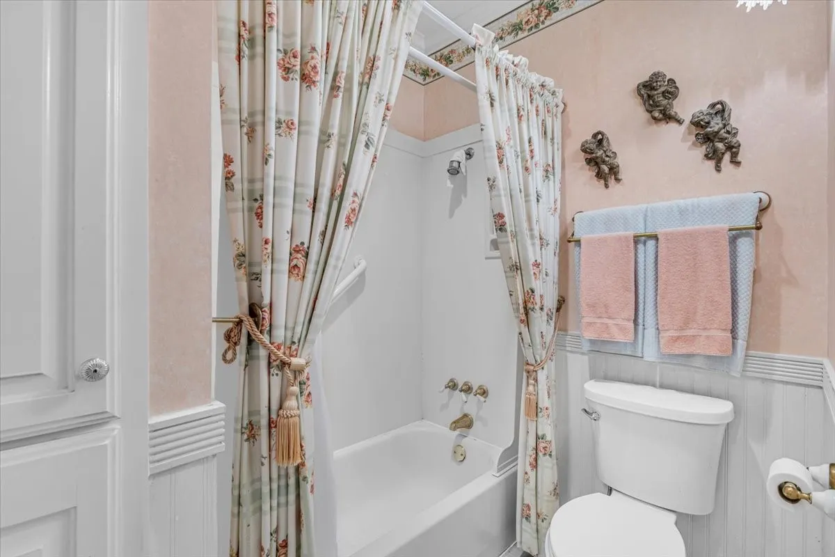 Built-in storage cabinet, full bathtub with shower, white commode, and wainscoting