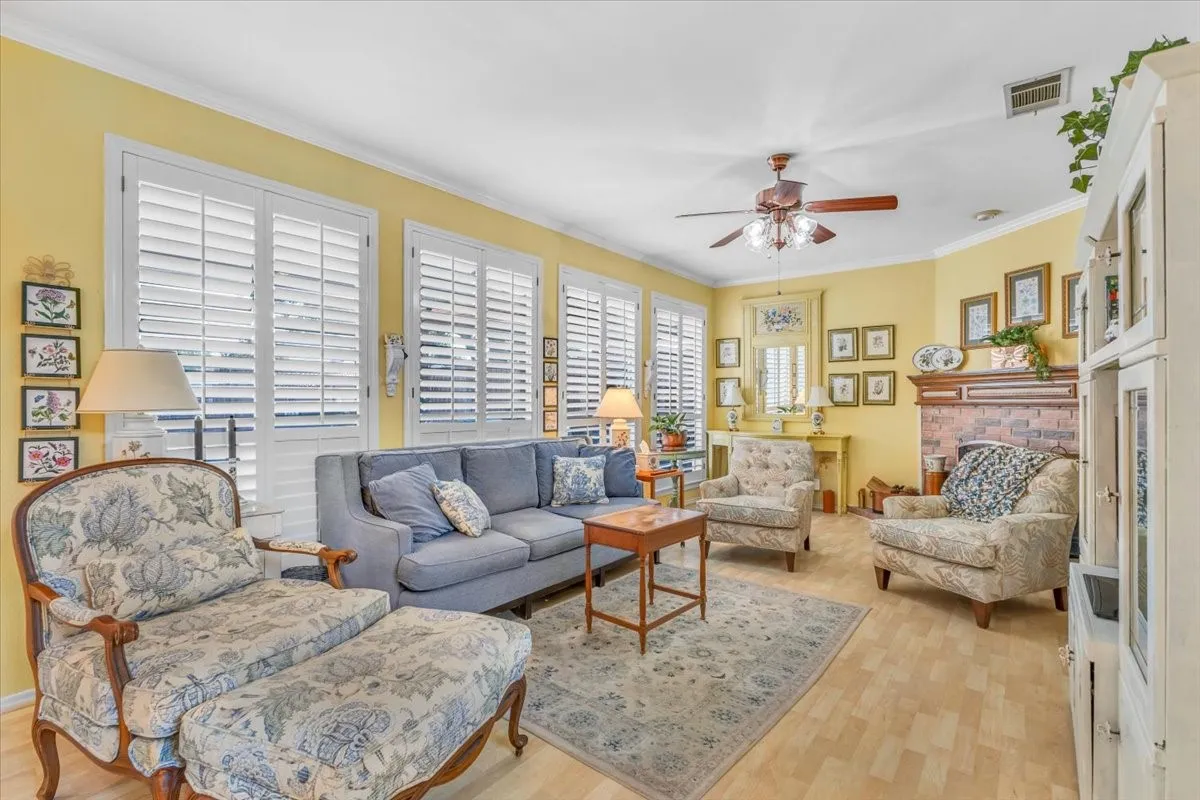 Living area featuring wood-finish flooring, multiple windows with white plantation shutters, a ceiling fan, and a brick fireplace with a wood mantel