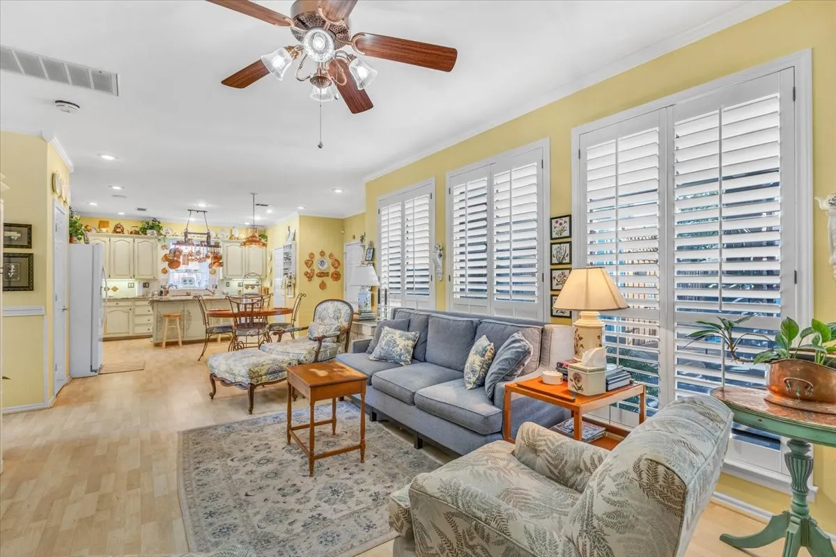 Open-concept living area with wood-finish flooring, a ceiling fan with integrated lighting, and multiple windows featuring plantation shutters