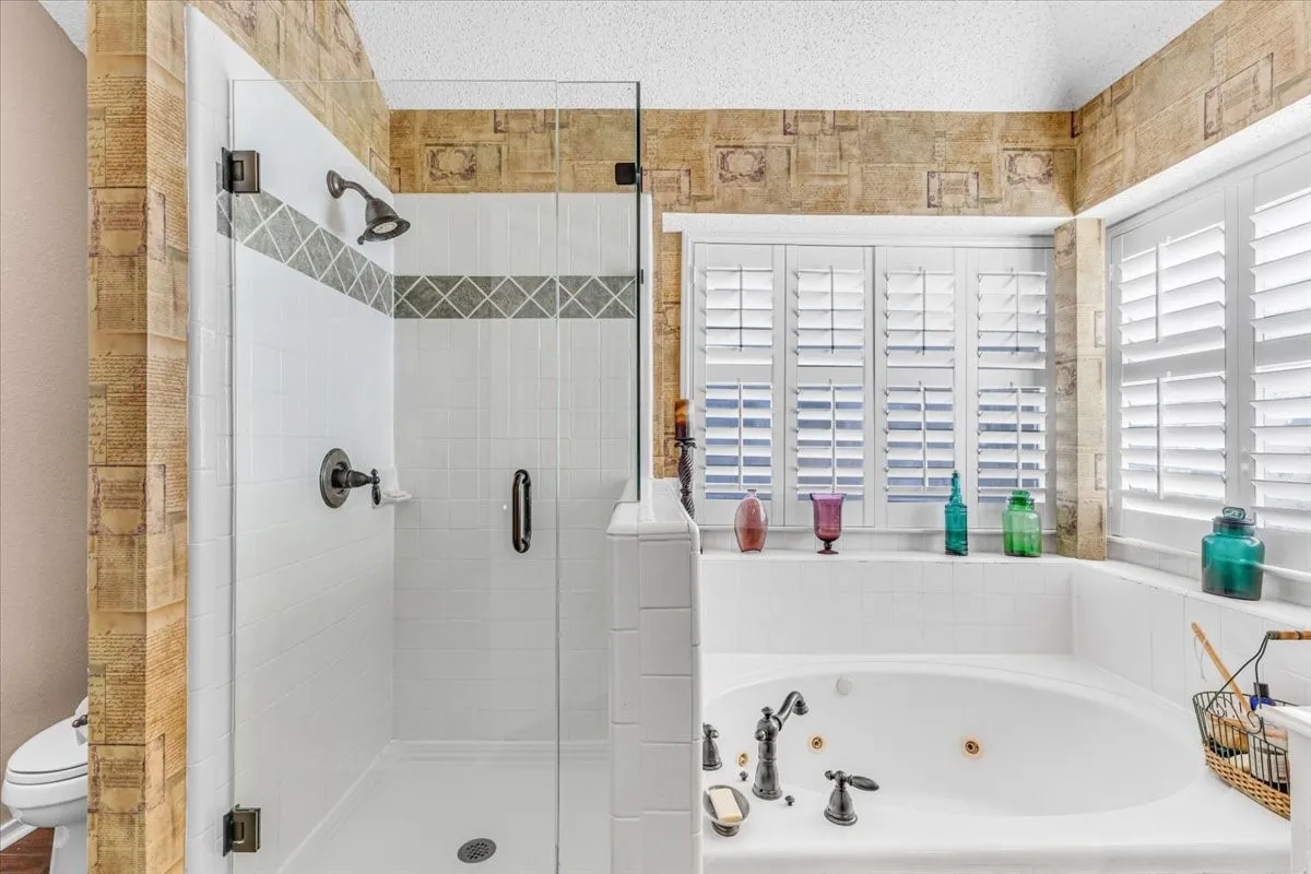 Glass-enclosed shower with a decorative tile band, soaking tub with dark bronze fixtures, and plantation shutters