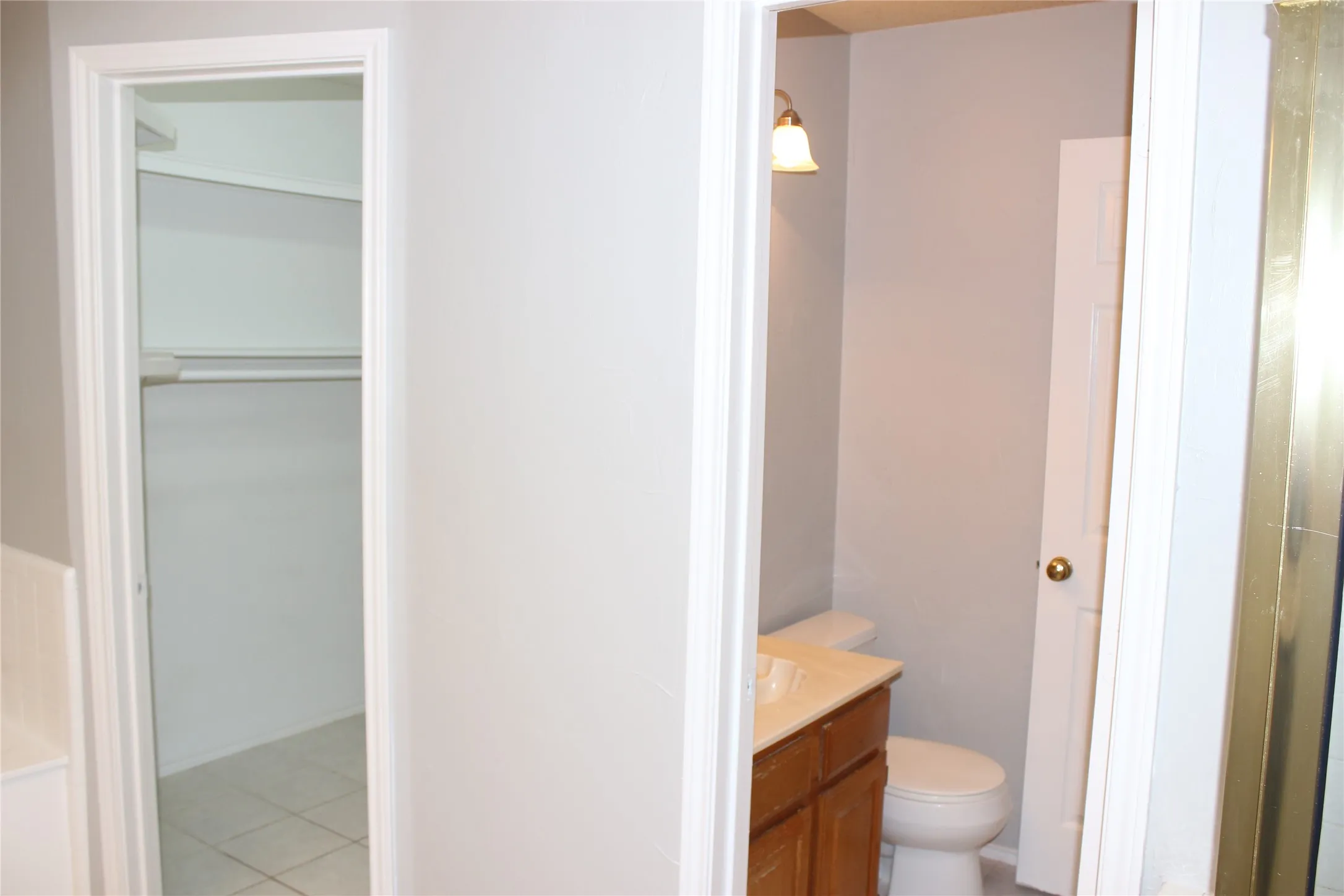 Interior view of a closet with white shelving and hanging rod, alongside a bathroom featuring a vanity with a light-toned countertop, a toilet, and a brass-toned wall sconce