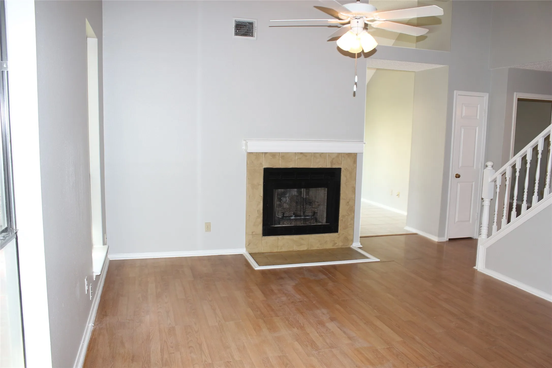 Spacious living area featuring a fireplace with a tiled surround, wood-finish flooring, a ceiling fan, and a staircase with a white railing