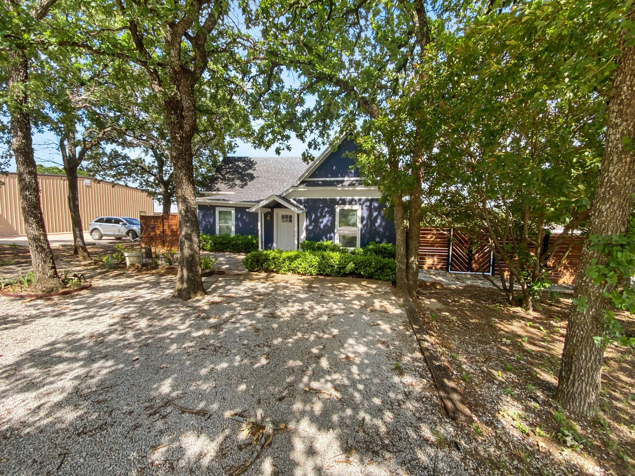 Deep blue exterior with white trim, covered front entry, gravel driveway, mature trees, and privacy fencing