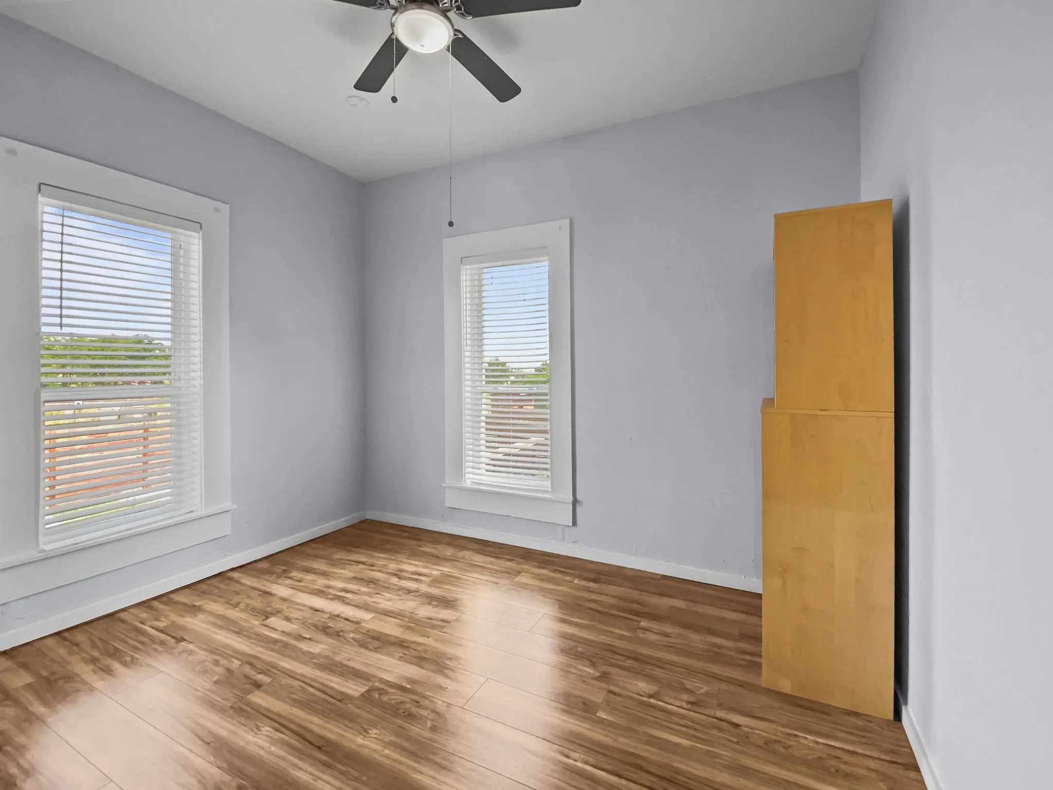 Wood-finish flooring, two windows with blinds, ceiling fan, and light blue wall color