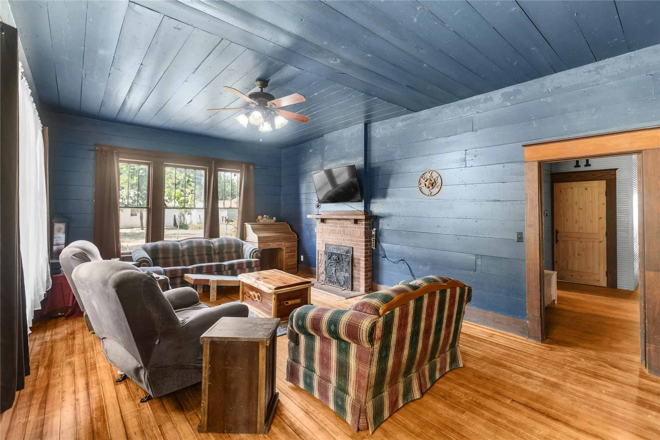 Hardwood flooring, painted wood plank walls, brick fireplace with iron grate, painted wood plank ceiling, and ceiling fan with light kit